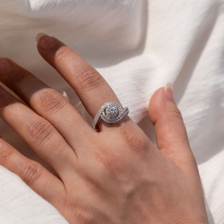 A Minifora Celestial Harmony diamond ring elegantly presented on a hand against a soft white fabric background. The ring features a round lab-created diamond at the center, surrounded by a double halo of smaller diamonds, with a split shank pavé setting, all beautifully crafted in 14K white gold.