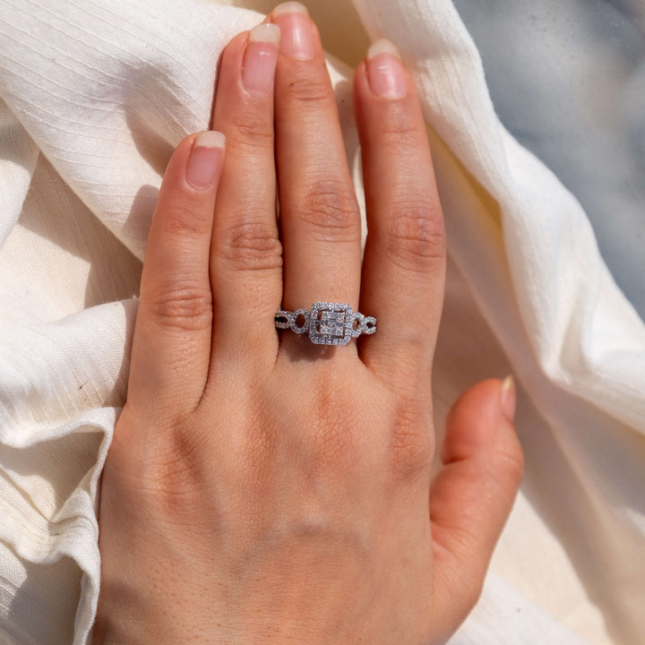 A hand is elegantly displaying Minifora's 'Elegant Radiance' engagement ring. The ring features a cushion-cut lab-created diamond at the center, framed by a halo of smaller diamonds, with two additional diamonds set on the band to create a symmetrical, eye-catching look. The entire piece is expertly crafted in 14K white gold.