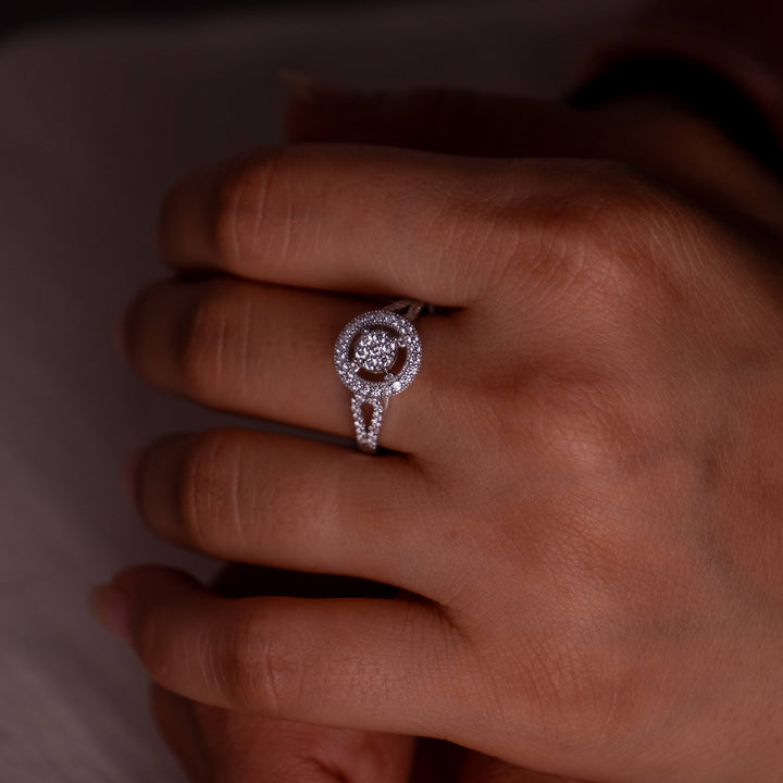 An Elysian Halo Ring from Minifora elegantly adorns a finger, its intricate design highlighted against a subdued background. The ring features a central round lab-created diamond encased in a halo setting, with additional diamonds decorating the loop on the band, all meticulously set in polished 14K white gold for a captivating sparkle.