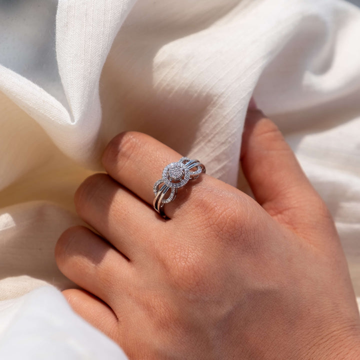 A hand is delicately framed by sheer white fabric, showcasing the Eternal Embrace Diamond Ring from Minifora. This stunning piece features intertwining bands of 14K white gold, each band sparkling with pavé-set lab diamonds that gracefully encircle a central round diamond halo. 