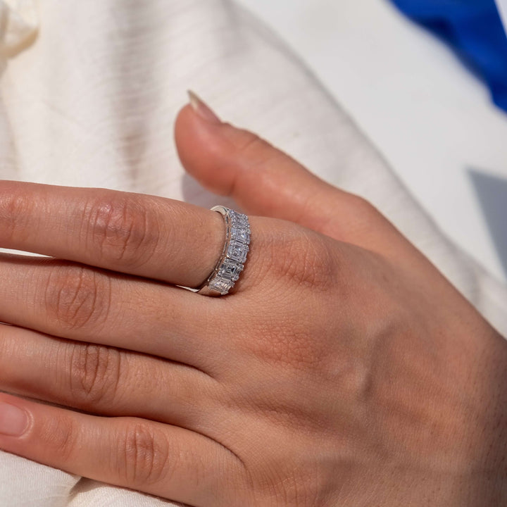 A finger is adorned with the Eternal Glitter Band from Minifora, captured against a backdrop of soft white fabric. This band, crafted in gleaming 14K white gold, boasts a series of baguette-cut lab-created diamonds in a channel setting, interspersed with rounds of pavé diamonds.