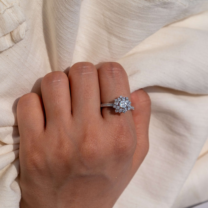 A delicate hand is tucked into the folds of a cream-colored, gauzy fabric, drawing focus to a Minifora 'Lumina Blossom' ring. This stunning piece features a cluster of lab-grown diamonds arranged in a floral pattern at the center, with the petals made up of diamonds, all set in a classic 14k white gold band. 