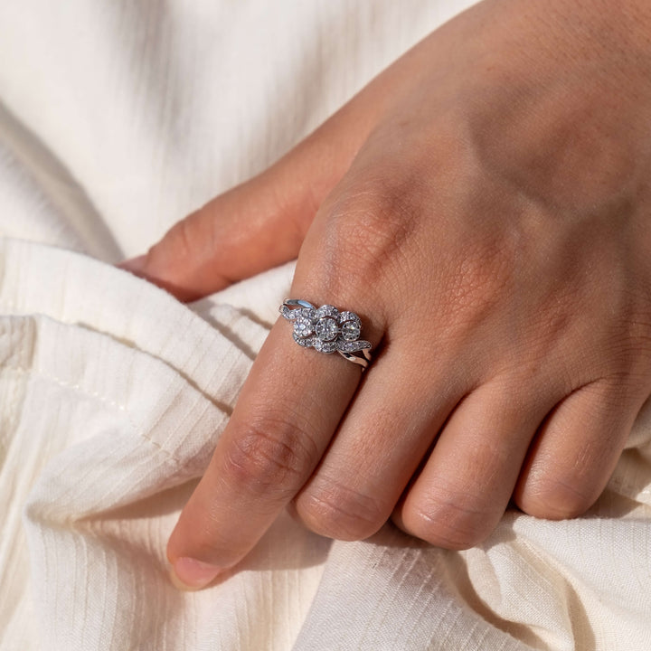 A gently rested hand against a light, textured fabric backdrop features a Minifora 'Radiant Embrace' ring. This exquisite ring boasts a floral-inspired design with lab-grown diamonds forming a radiant cluster, embraced by a split shank band of 14k white gold that intertwines elegantly.