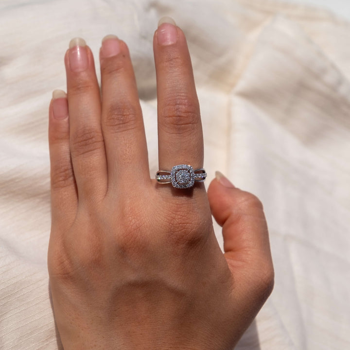 A hand is gracefully positioned against a backdrop of natural linen, highlighting a striking Minifora 'Radiant Harmony' ring. This ring features a square-cut lab-grown diamond, framed by a glittering halo and set upon a band of 14k white gold. The band itself is adorned with smaller diamonds along its shoulders.